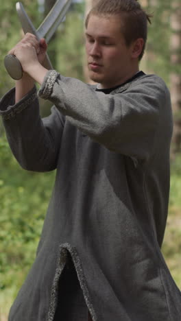 young man on loose long shirt does sword strikes training for battle in sunny wood slow motion. medieval soldier with armor. alternative sports camp