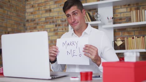 caucasian man making video call using laptop holding handwritten sign making marriage proposal