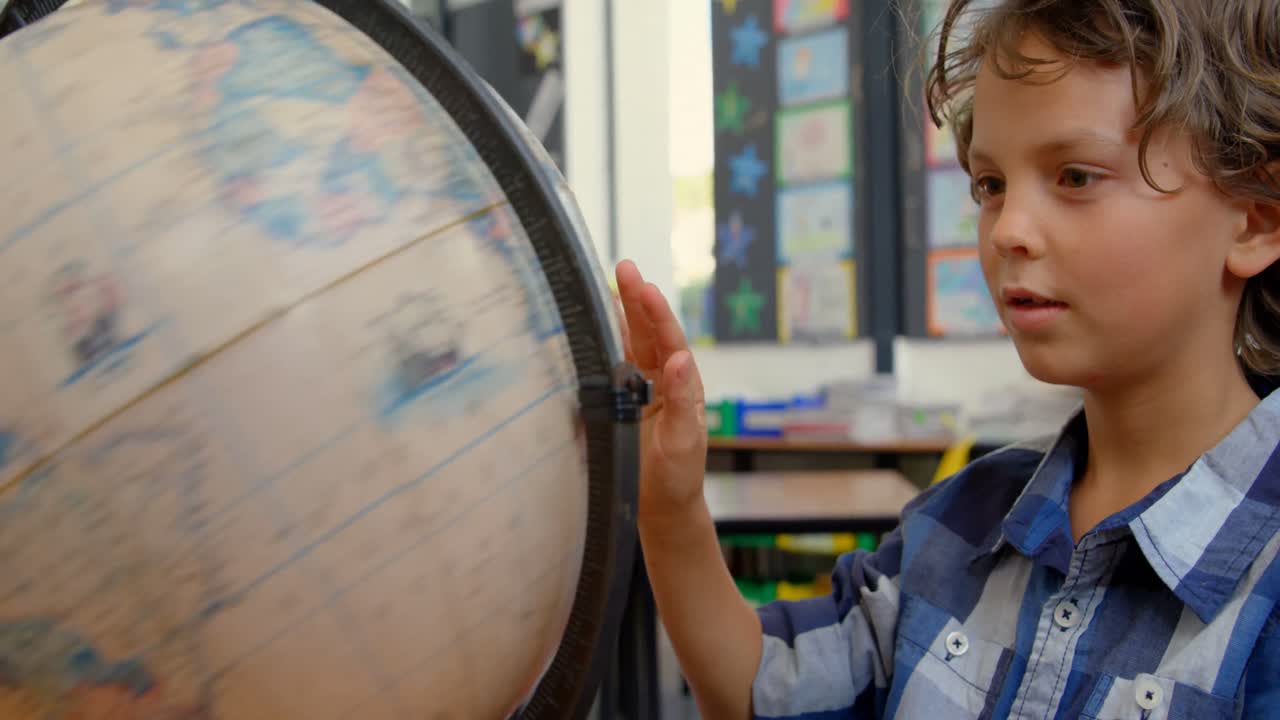 vista frontal de un escolar caucásico estudiando el globo en el escritorio en el aula de la escuela 4k