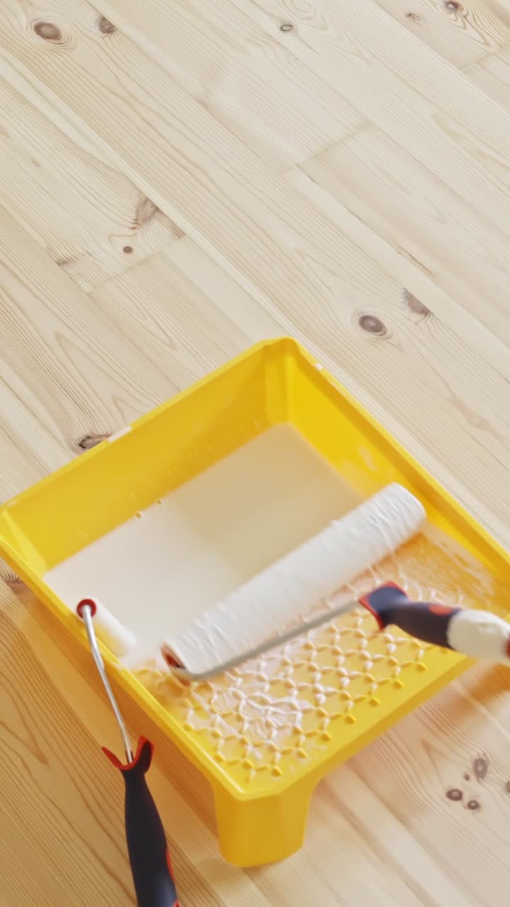 A vertical close-up captures a white paint roller resting in a bright yellow tray filled with white paint, placed on a natural wooden floor during indoor wall renovation and preparation work