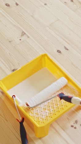 a vertical close-up captures a white paint roller resting in a bright yellow tray filled with white paint, placed on a natural wooden floor during indoor wall renovation and preparation work