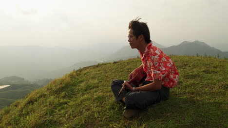 Close-up-shot-of-an-Asian-man-hiker-sitting-on-grass-on-top-on-a-mountain-and-playing-ukulele-on-a-windy-morning