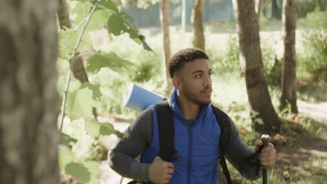 african american man wearing backpack and hiking with trekking poles in forest, slow motion