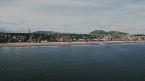 Establishing-aerial-shot-of-the-North-Sea-and-Portobello-in-Edinburgh,-with-low-tide-beach,-residential-buildings-a-church-and-Arthur's-Seat-in-the-background