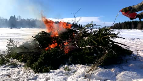 Zeitlupe-Von-Lagerfeuer,-Kiefernholz-Und-Fichtenzweigen,-Die-Auf-Dem-Verschneiten-Eis-Brennen,-Winter,-Mann,-Der-Dem-Feuer-Öl-Hinzufügt