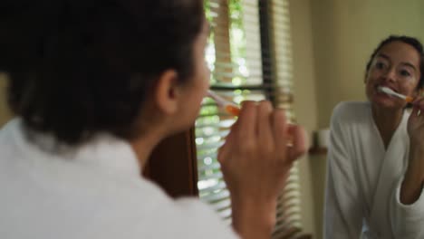 Back-view-of-biracial-woman-with-vitiligo-wearing-robe-looking-in-mirror-and-brushing-teeth