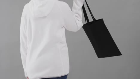 caucasian woman wearing white hoodie holding black bag on grey background, copy space, slow motion