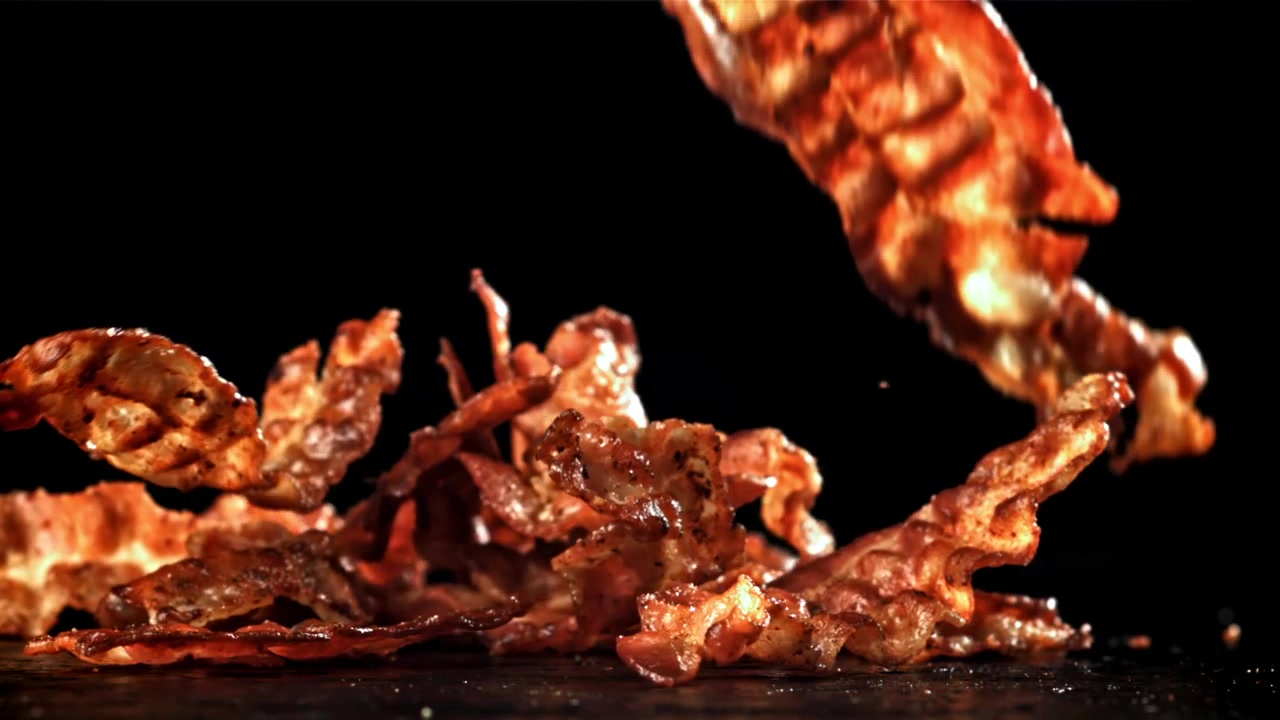 Sprøde stegte baconstrimler falder i slow motion på sort baggrund