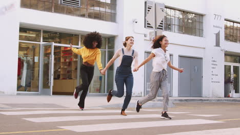 three millennial girlfriends holding hands and laughing as they run across a pedestrian crossing