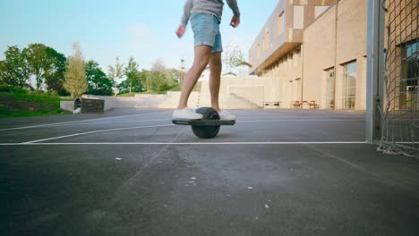 young mand boarding one wheel skating away in schoolyard with threes and blue sky during summer shot from frogs eye perspective