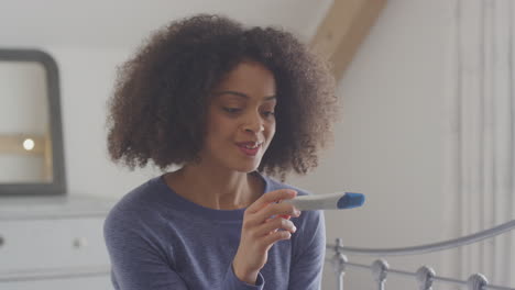 excited woman sitting on bed in bedroom with positive home pregnancy test