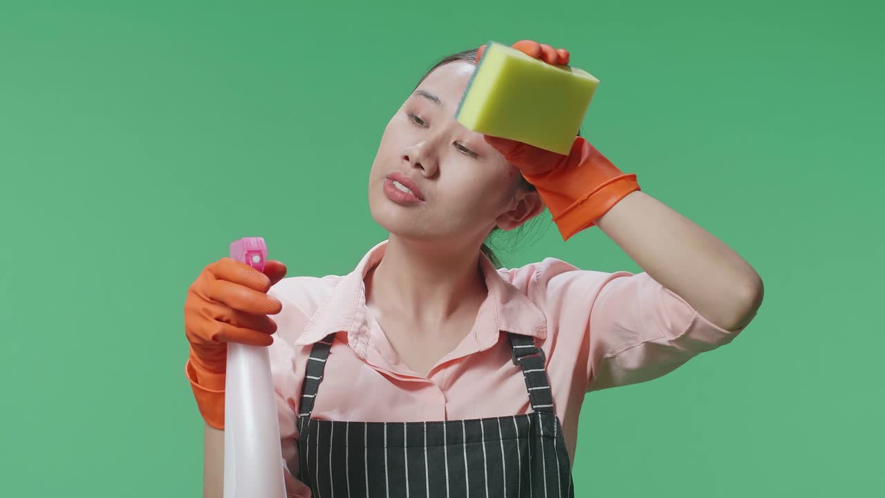피곤한 아시아 여성 집주인이 스프레이와 부 스폰지를 들고 초록색 스크린 스튜디오에서 을 고 있습니다.