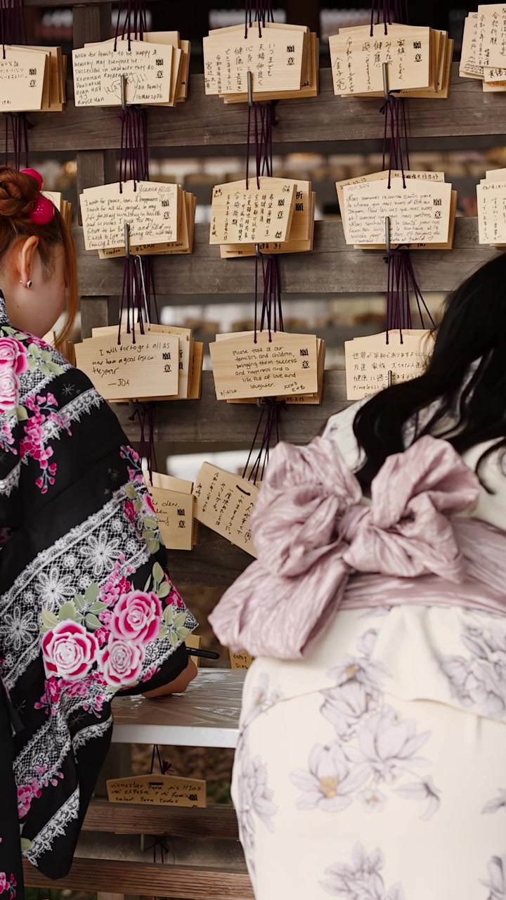 Rear view of women in floral kimonos writing wishes on traditional wooden plaques in Tokyo.