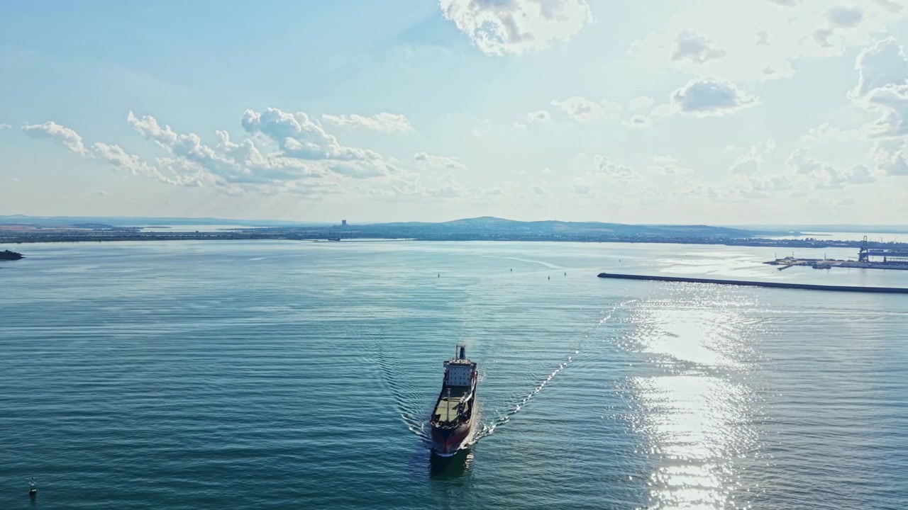 Grande nave da carico naviga in acque calme sotto un cielo soleggiato in un'area portuale a mezzogiorno.