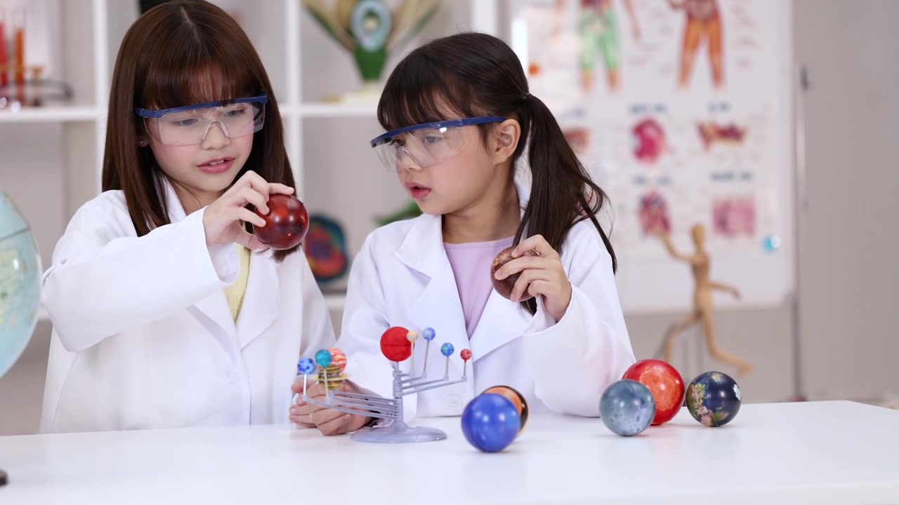 Dua gadis mengenakan kacamata pengaman dan jas lab memeriksa model tata surya di ruang kelas