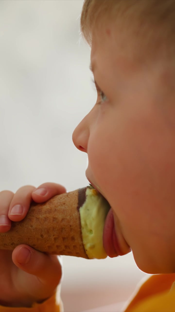 Primo piano di un bambino piccolo che mangia un cono gelato. La sequenza mostra la progressione del leccare e consumare gelato verde dalla cialda tenuta in mano, con una manica gialla visibile. Illuminazione soffusa.