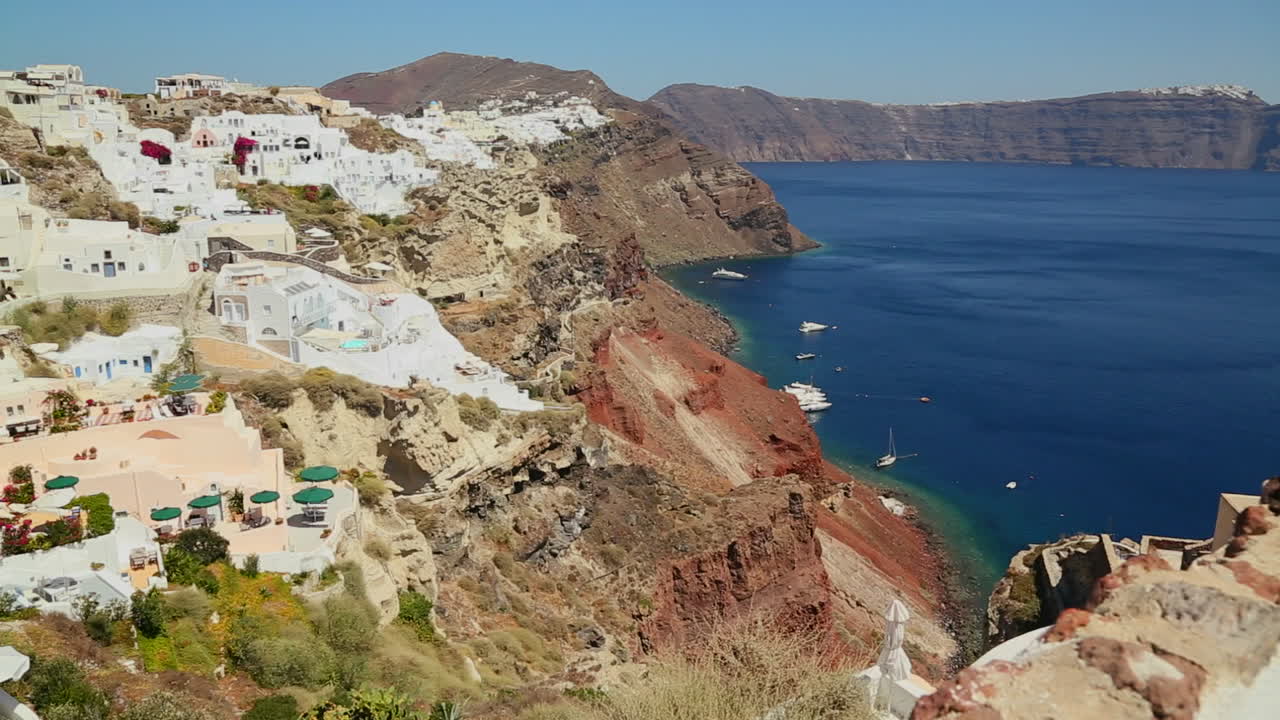 panorámica lenta de casas blancas que bordean las laderas de la isla griega de santorini con una bandera griega en la distancia