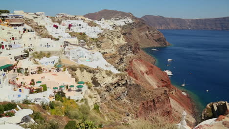panorámica lenta de casas blancas que bordean las laderas de la isla griega de santorini con una bandera griega en la distancia