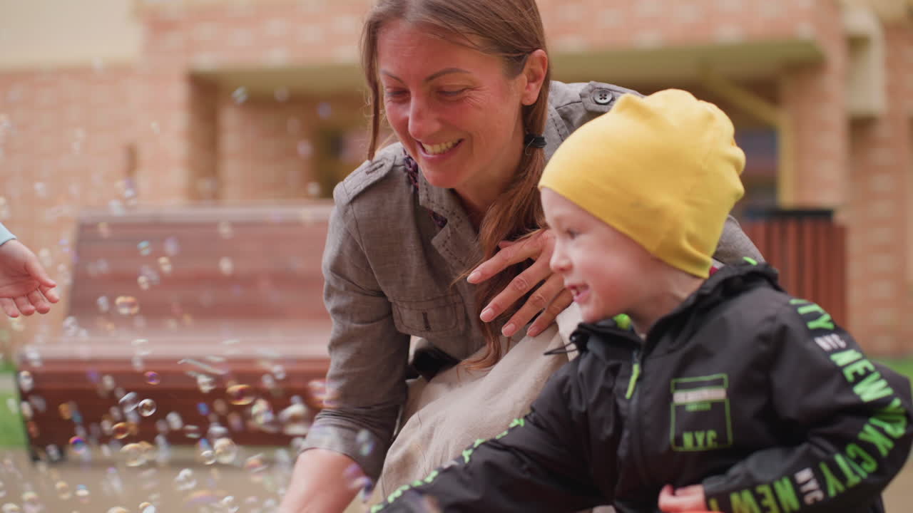 Happy mother bends close watching her son in yellow cap joyfully playing with floating soap bubbles on sunny courtyard, sharing laughter and bonding during cheerful outdoor activity