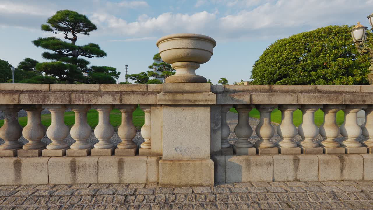 A close-up shot of a classical stone balustrade with ornamental urns and manicured Japanese pines in the background