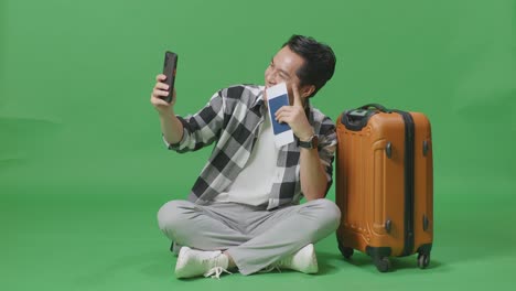 full body of asian male traveler with luggage and passport smiling and taking photo on smartphone while sitting in the green screen background studio