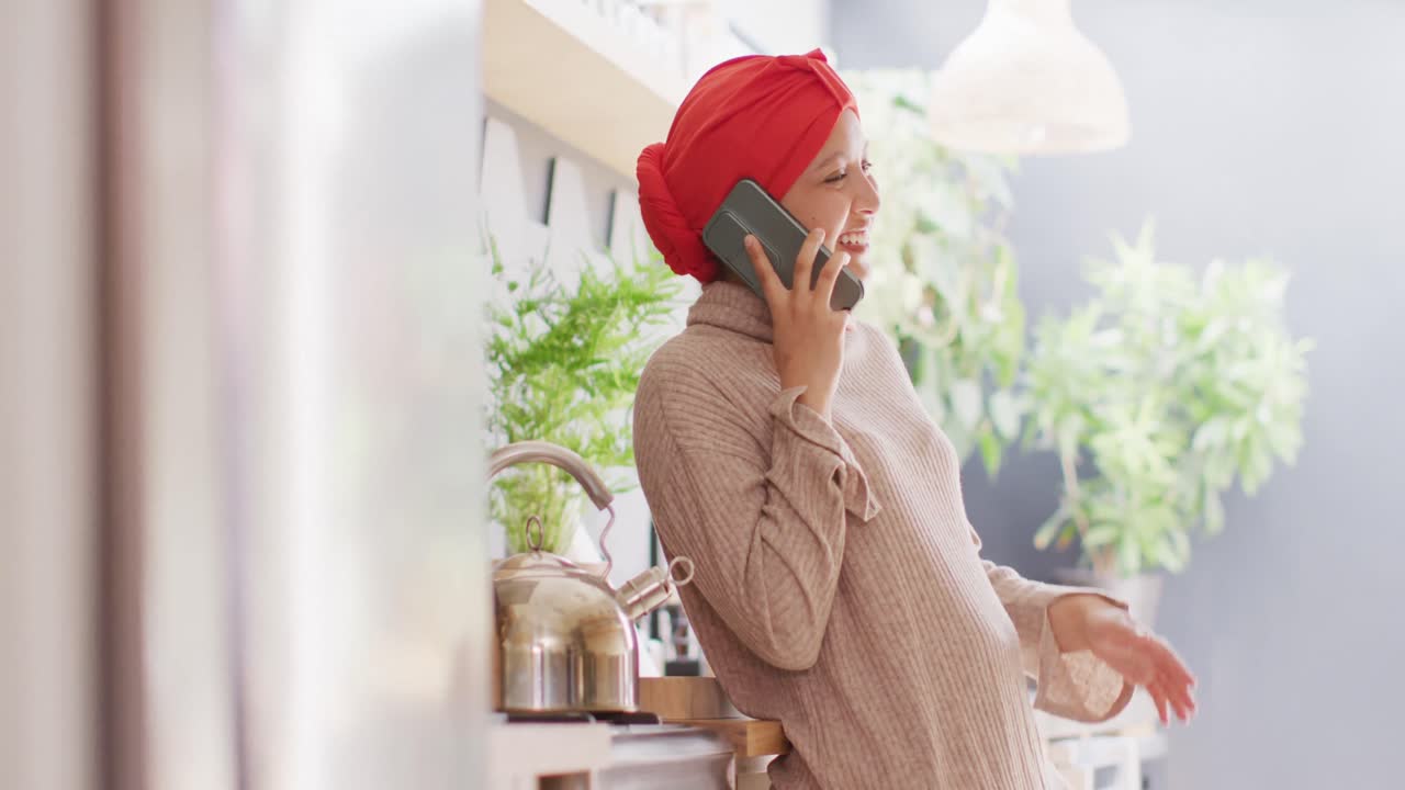 Video of laughing biracial woman in hijab talking on smartphone standing in kitchen at home