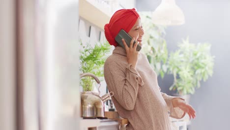 video de una mujer biracial riendo con hijab hablando en un teléfono inteligente de pie en la cocina de su casa
