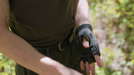 adventurer pulling off wool fingerless glove, hairy forearm and wrist revealed, belt and cargo pants visible, moving hands in sunlit forest clearing with blurred foliage background swaying