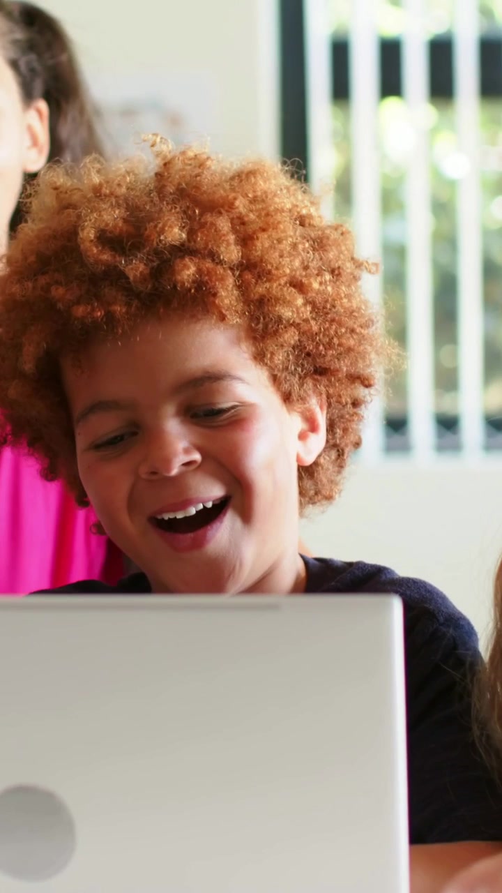 Vertical video: Clip starting, curly redhead boy watching laptop, girl in pink leaning, laughing
