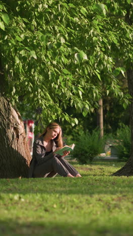 lady sitting outdoors, under a tree on grassy field, reading book with head tilted slightly under warm sunlight, tree leaves sway gently in breeze, with background featuring residential building