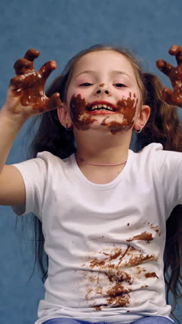 cheerful little girl licking chocolate from her fingers. dirty face and white t-shirt of a happy child. funny kid in smeared chocolate isolated on blue. vertical video