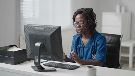 african female medical assistant wears white coat headset video calling distant patient on computer. doctor talking to client using virtual chat computer app. telemedicine remote healthcare