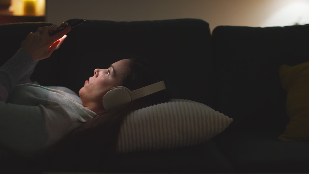 무선 헤드폰을 착용 한 여성이 밤에 집에서 소파에 누워서 휴대 전화로 온라인 콘텐츠를 스트리밍하거나보고 있습니다.