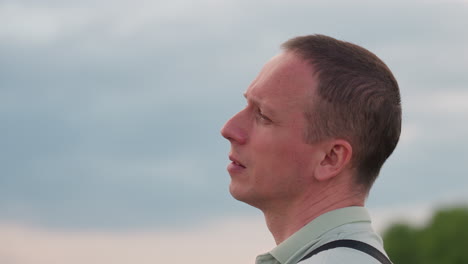 closeup side profile of man gazing into distance with thoughtful expression wearing light grey polo shirt and shoulder strap while standing near parked vehicle against blurred field under cloudy sky