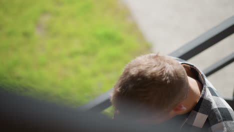 top down view of student in black white plaid shirt retrieving cigarette from ear flicking lighter ignites tip sparking bright ember against blurred greenery and metal railing in sunlit scene