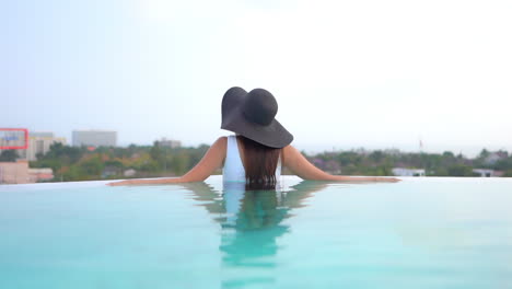 vista trasera de una mujer elegante con sombrero de verano flexible y traje de baño en la piscina con el paisaje urbano de fondo