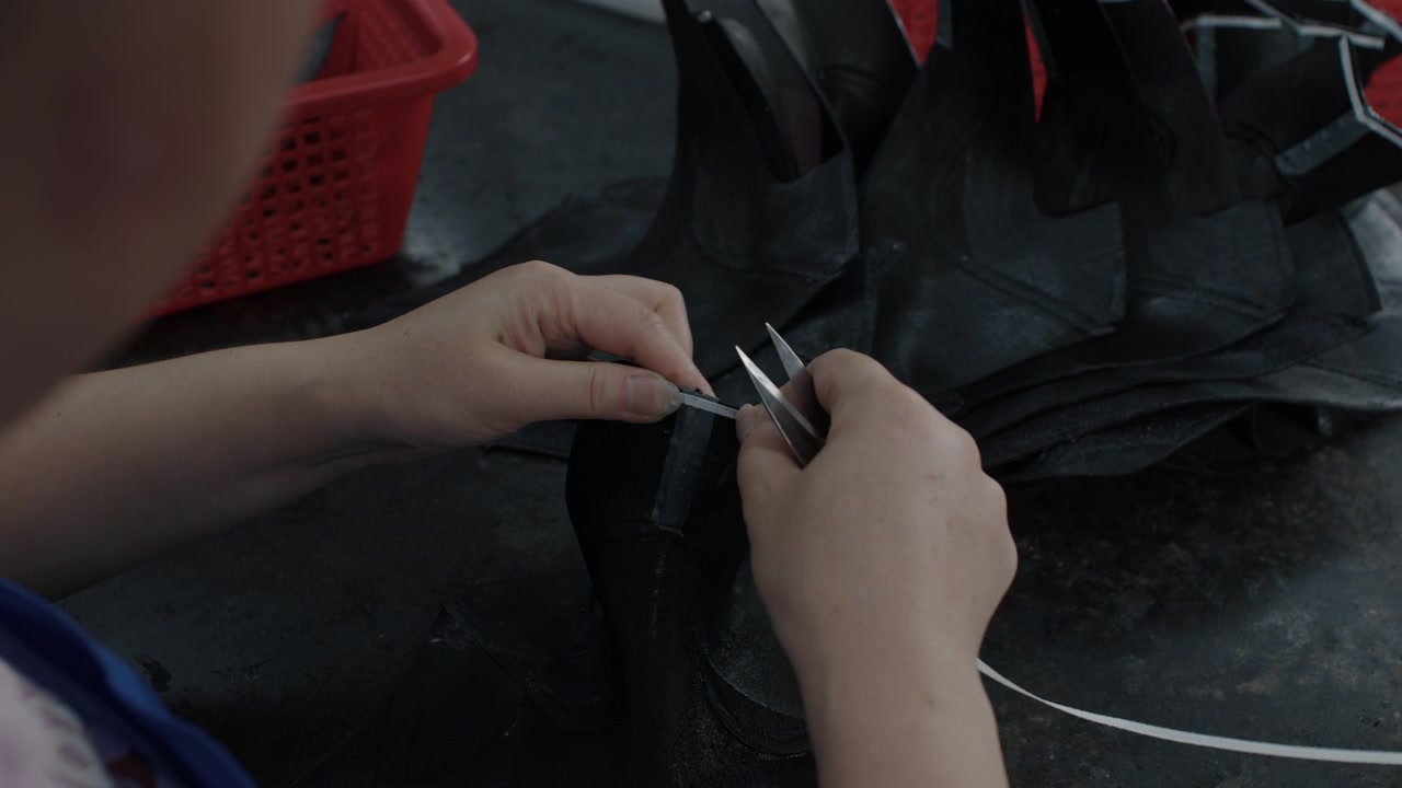 Mano de trabajador recorta las partes superiores de zapatos negros en una mesa de fábrica con cestas rojas cerca.