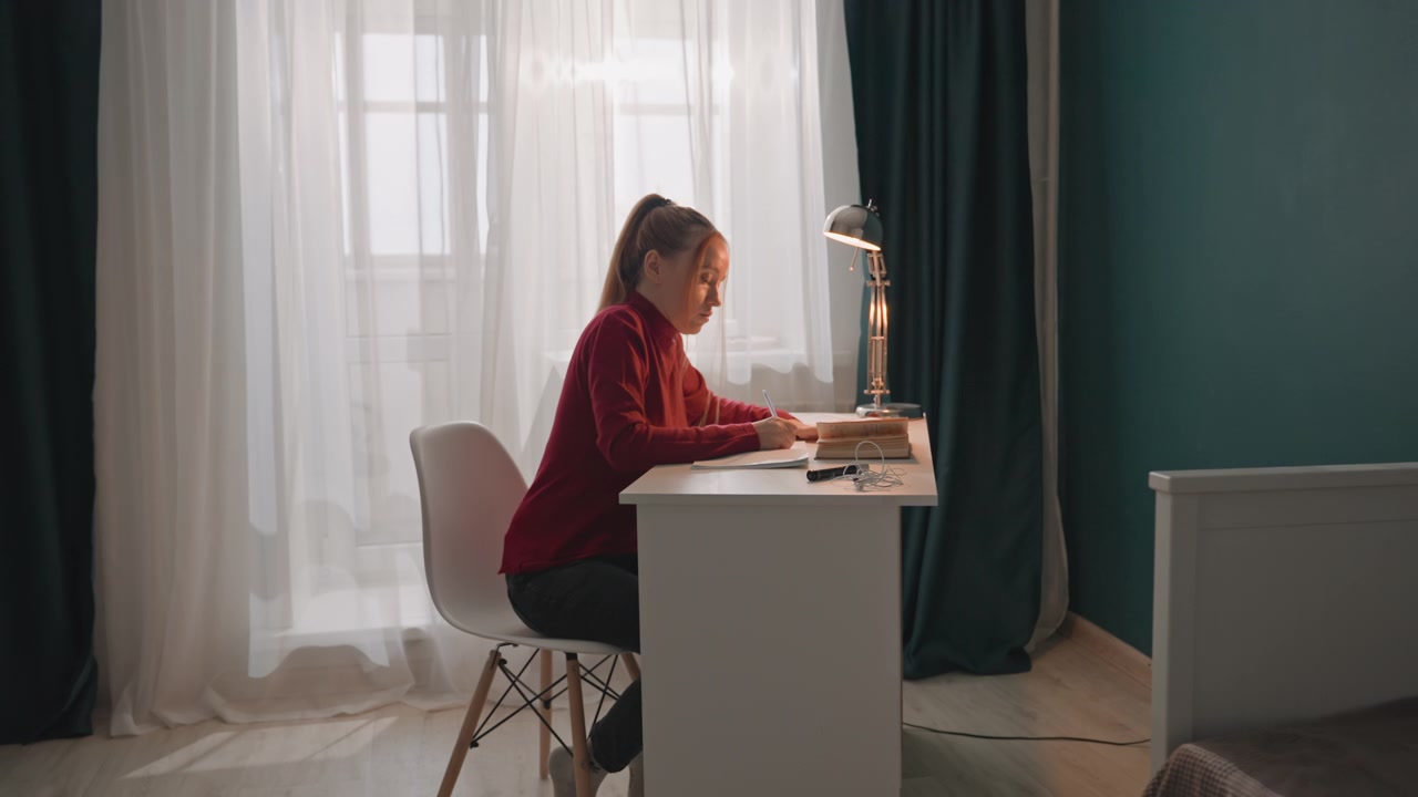 Mladá žena v červeném topu sedí u bílého stolu a pracuje. Místnost má tyrkysové stěny s tmavě zelenými závěsy a přirozeným světlem. Kovová lampa osvětluje pracovní prostor, zatímco žena pracuje.