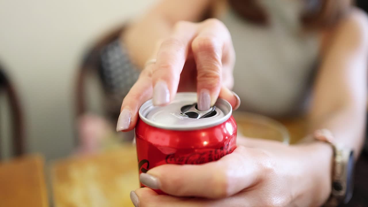 A woman’s hands open a red soda can on a wooden table in soft natural light, captured in a steady close-up with shallow depth of field