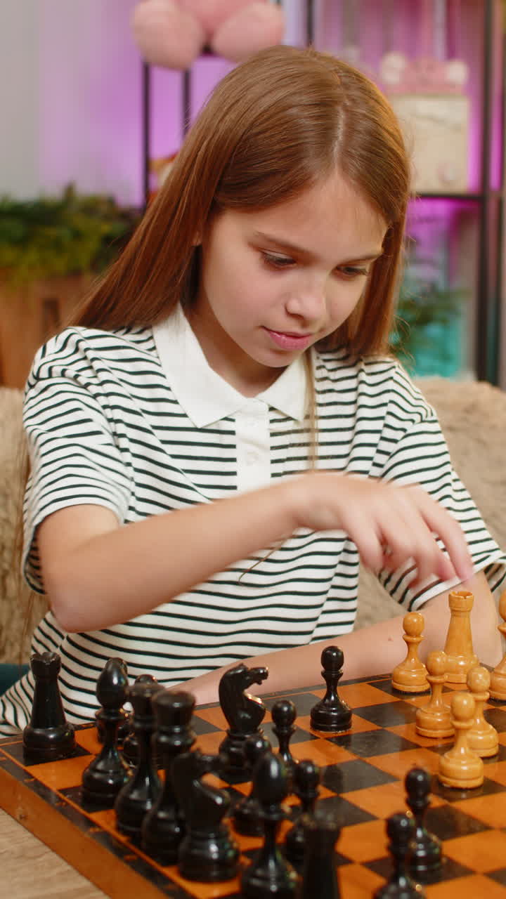 Child girl kid playing chess alone at home focused on move sitting at table on home sofa indoors