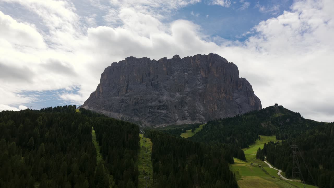 Sassolungo mountain group, peaks rising above pine forest and green meadows, Dolomites, Italy. Aerial drone lateral view