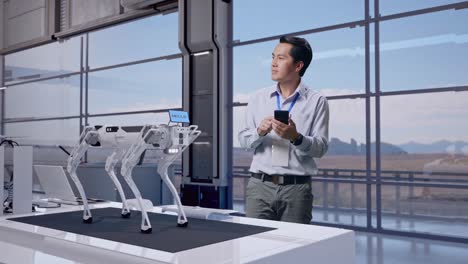 full body of an asian male professional worker standing with his smartphone in high tech factory, robot dog, he observes by looking around before he come to concentrating with his smartphone