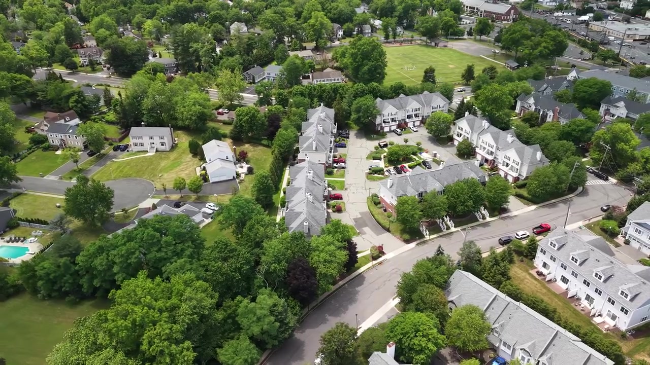 High aerial view flying over a typical american suburban neighborhood with townhouse developments, lush green trees, quiet streets, and parked cars on a sunny day in a small town