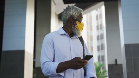 African-american-senior-man-wearing-face-mask-using-smartphone-and-looking-around-in-corporate-park