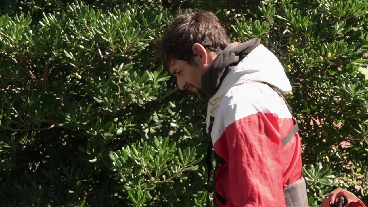 Man wearing a red jacket maintains his garden by cutting weeds and grass with a manual string trimmer