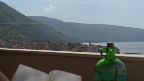 Woman-reading-book-with-view-of-seaside-town,-rising-shot,-Komiza,-Vis,-Croatia