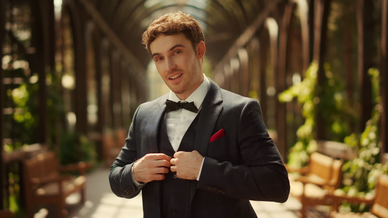 정원에서 아치 아래 포즈를 취하는 매력적인 신랑. 공원에서 재 ⁇ 을 버튼하는 남자