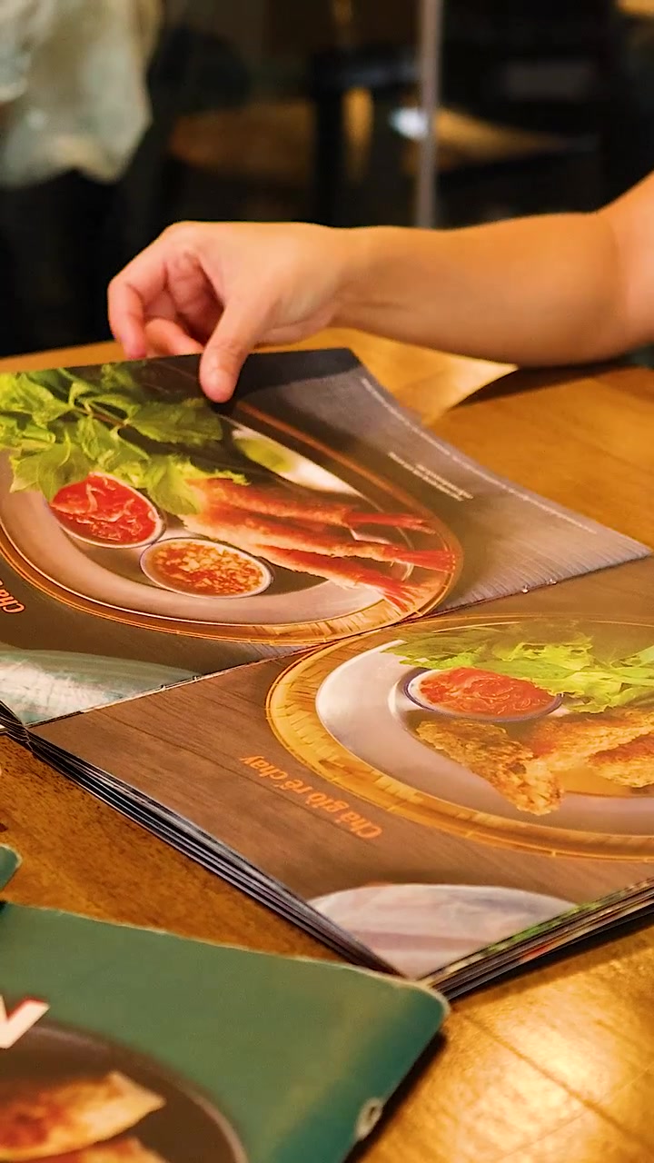 En person bladrer igennem en vietnamesisk restaurantmenu, der viser farverige retter under varm belysning på et træbord