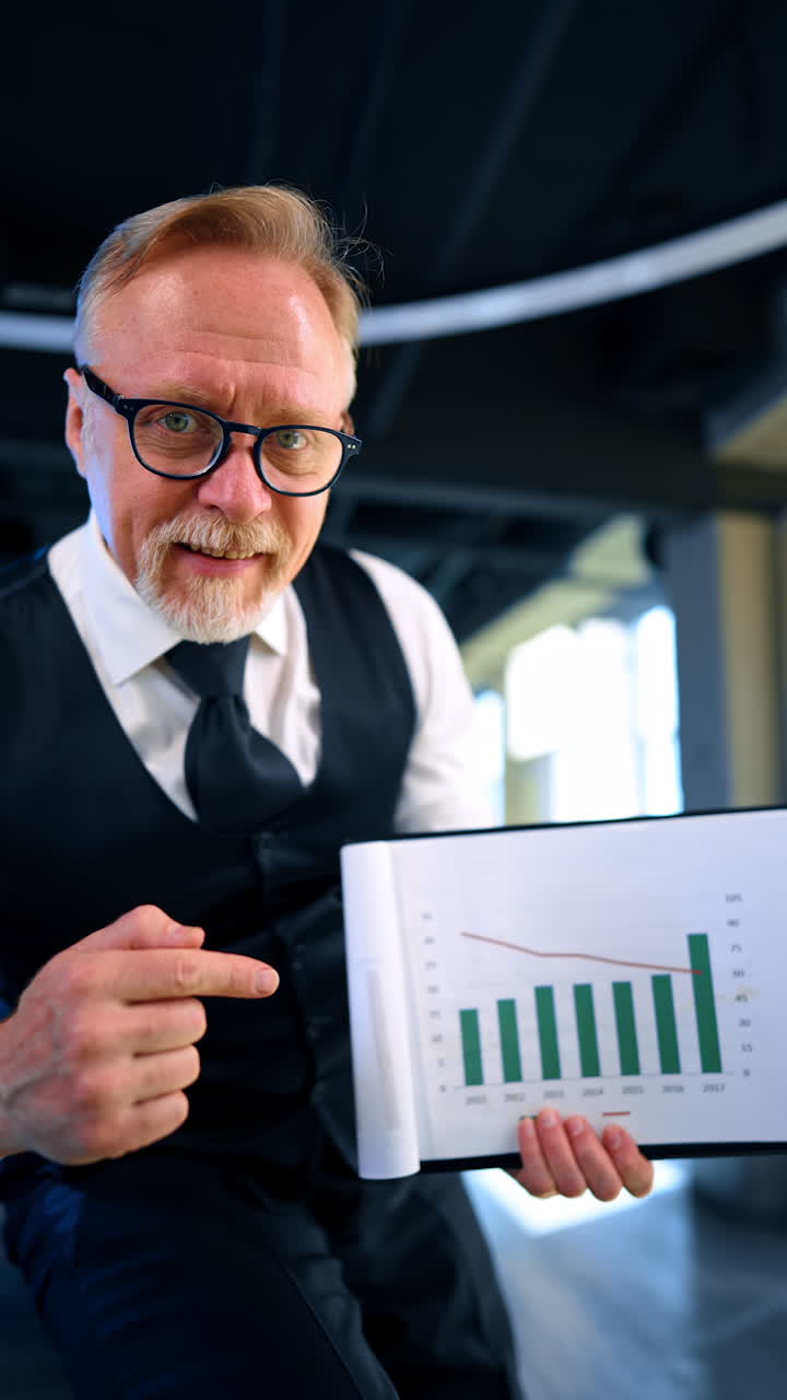 Excited positive man in official clothes and glasses showing the chart to the camera. Businessman happy about work analysis results. Vertical video.