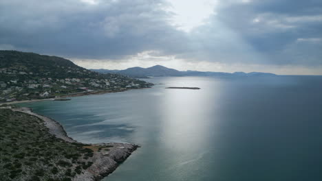 a drone above the athens coastline, capturing the stunning reflection of sunlight on blue waters. the sky is overcast, with sunlight breaking through clouds, revealing a coastline lush with greenery.